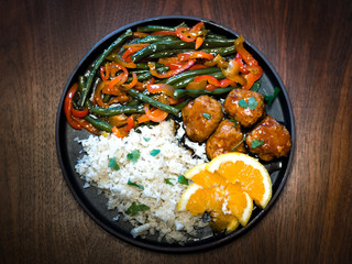 Close up photograph of a low carb meatball dinner with side of mashed cauliflower and string beans and red pepper garnished with orange slices and cilantro served on a black plate with wood background