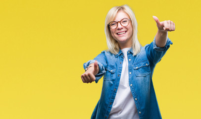 Young beautiful blonde woman wearing glasses over isolated background approving doing positive gesture with hand, thumbs up smiling and happy for success. Looking at the camera, winner gesture.