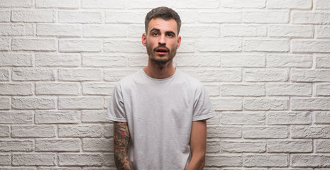 Young adult man standing over white brick wall scared in shock with a surprise face, afraid and excited with fear expression