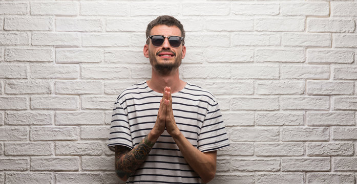 Young adult man wearing sunglasses standing over white brick wall begging and praying with hands together with hope expression on face very emotional and worried. Asking for forgiveness