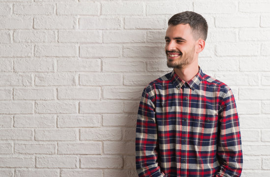 Young adult man standing over white brick wall looking away to side with smile on face, natural expression. Laughing confident.