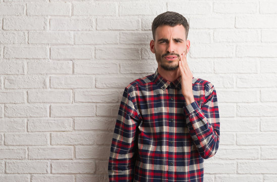 Young adult man standing over white brick wall touching mouth with hand with painful expression because of toothache or dental illness on teeth. Dentist concept.