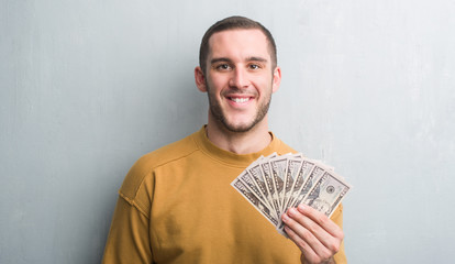 Young caucasian man over grey grunge wall holding dollars with a happy face standing and smiling...