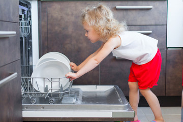 Cute blonde toddler girl helping in the kitchen taking plates out of dish washing machine