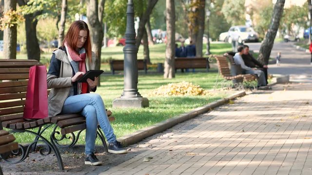 Modern woman with red hair sitting on a bench and using the tab. Early autumn