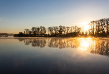 Obraz premium Autumn forest is reflected in the mirror of the river, covered with remnants of fog, which is scattered under the rays of the rising sun.