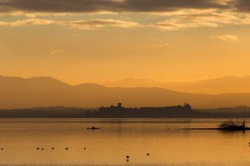 Obraz na płótnie Canvas Beautiful view of Trasimeno lake (Umbria, Italy) at sunset, with orange tones, birds on water, a man on a canoe and Castiglione del Lago town on the background