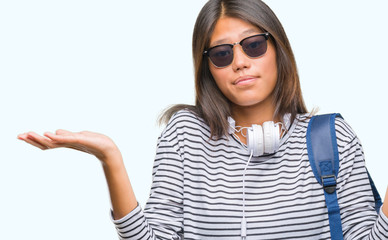 Young asian student woman wearing headphones and backpack over isolated background clueless and confused expression with arms and hands raised. Doubt concept.