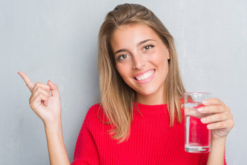Beautiful young woman over grunge grey wall drinking a glass of water very happy pointing with hand...