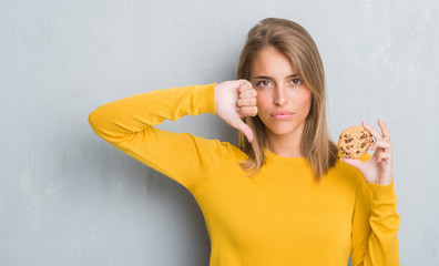 Beautiful young woman over grunge grey wall eating chocolate chip cooky with angry face, negative...