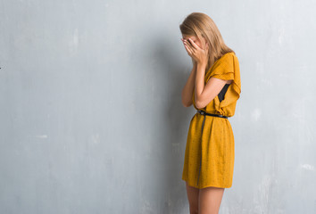 Beautiful young woman standing over grunge grey wall wearing a dress with sad expression covering face with hands while crying. Depression concept.