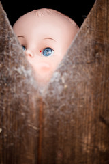 Old plastic doll with bright blue eyes looking through a gap in a wooden panel door. The gap is...