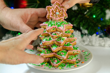 Human hands reach for the pieces gingerbread tree on the background of the Christmas tree, on which lights festooned.