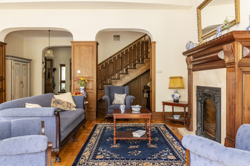 A luxurious living room interior with antique sofa and coffee table in front of a fireplace in an English style mansion. Wooden staircase in the background. Real photo.