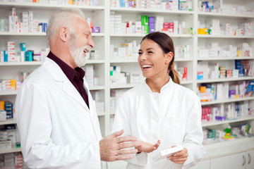 Obraz premium Medicine, pharmaceutics, health care and people concept - Young female and senior male pharmacists smiling and standing in front of shelves with medications