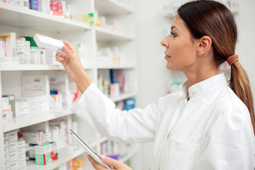 Beautiful young female pharmacist reaching for medications on the shelf, holding a tablet in one hand
