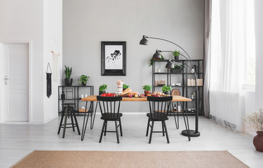 Black chairs at dining table with food in apartment interior with lamp and poster on grey wall....