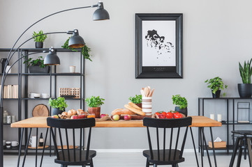 Lamp above black chairs and wooden table with food in grey dining room interior with poster. Real...