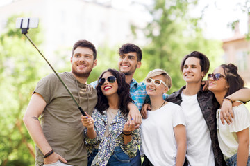 friendship, leisure and technology concept - group of happy smiling friends taking picture by selfie stick at summer park