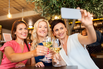 people, technology and lifestyle concept - women drinking wine and taking selfie by smartphone at bar or restaurant