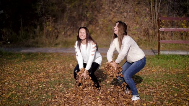Two beautiful woman throw golden leaves in autumn garden. Girls jump, have fun, act like children. 4k 60fps slow motion