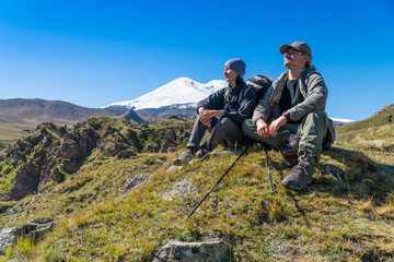 Man and woman with backpack admiring Elbrus mountain view landscape from the cliff edge. Enjoying nature vacation travel adventure at Caucasus mountains.