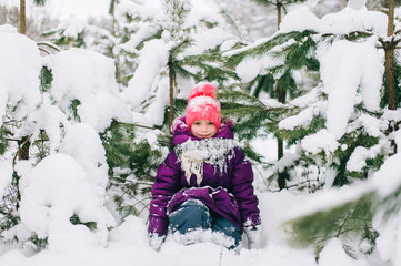 Adorable little girl posing under snowy spruce tree in wood outdoor. Beautiful female child sitting on snow in front of christmas tree in forest. Winter portrait of lovely young baby.  Funny kid face.