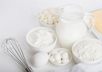 Fresh dairy products on white table background. Glass jar of milk, bowl of sour cream, cottage cheese and baking flour and mozzarella. Eggs and cheese. Steel whisk.