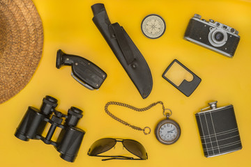 Traveler's things: hat, binoculars, glasses, compass, watch, knife, camera, flask on a yellow background.