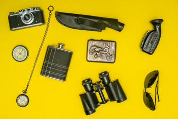 Traveler's things: hat, binoculars, glasses, compass, watch, knife, camera, flask on a yellow background.