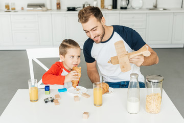 handsome young father and son playing with toy plane while having breakfast together at home