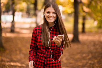 Pretty girl holding phone and listening music with her headphones in autumn