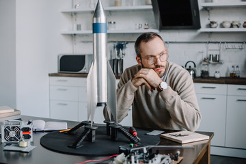 thoughtful engineer sitting at table with rocket model at home and looking away