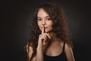 Body language. Portrait of beautiful charming young European female with black eyes and mysterious smile, holding index finger at her mouth, having big secret, saying Shh and looking at camera