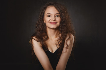 Femininity, beauty and body language. Isolated shot of cute pretty girl with curly dark hair and make up sitting indoors in closed posture and looking at camera with charming smile, feeling shy