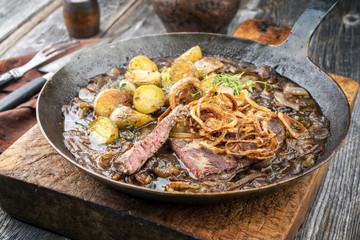Traditional dry aged sliced roast beef with fried onion rings and potato chips as closeup in a wrough-iron pan with brown sauce
