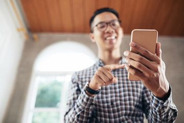 Low angle view of defocused young Asian man smiling cheerfully while taking selfie on modern smartphone