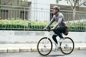Young Asian man wearing casual clothes, glasses and safety helmet riding bicycle along the road...