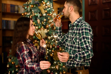 Merry Christmas and Happy New Year! Attractive young couple is celebrating holiday at home together, drinking champagne and smiling with Bengal lights in hand