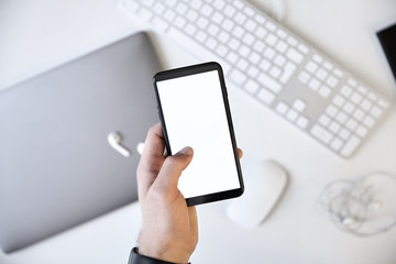 Left hand holding a white blank screen smartphone over the desk
