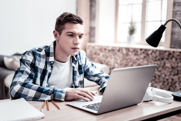 Modern laptop. Concentrated young student looking at the screen of his modern laptop while sitting in front of it at home