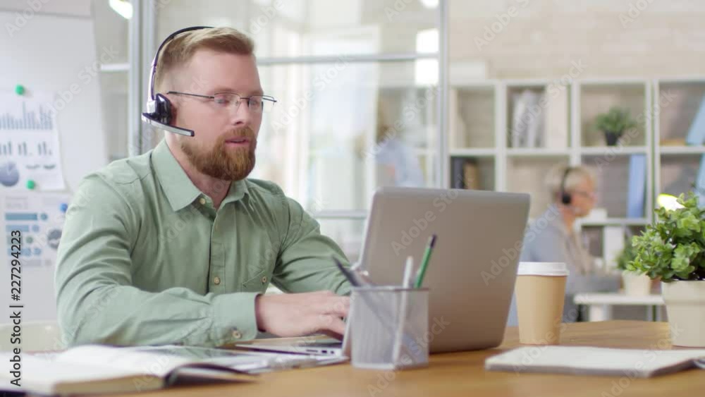Poster male call center manager in headset typing on laptop and talking via video call while working at des