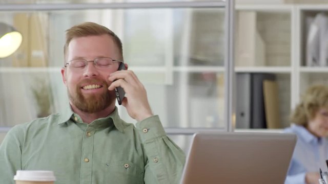 Joyous Caucasian office worker sitting at desk, smiling and chatting on mobile phone