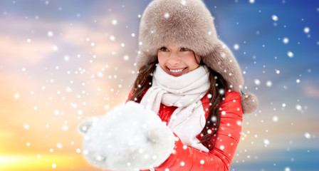 people, season and leisure concept - happy woman in winter fur hat holding snow in her hands outdoors over sky background and snow