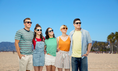 friendship, leisure and summer holidays concept - group of happy smiling friends in sunglasses hugging over venice beach background in california