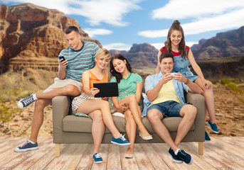 friendship, leisure and technology concept - group of happy smiling friends with tablet pc computer and smartphones sitting on sofa over grand canyon national park background