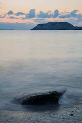 Long exposure capture with beach, stone and sunrise on coast of Mediterranean sea. In background a little island and cloudy sky.  