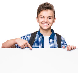 Portrait of handsome teen boy posing behind white panel isolated on white background. Teenager child holding empty billboard. Young student with school bag peeping behind blank board. 