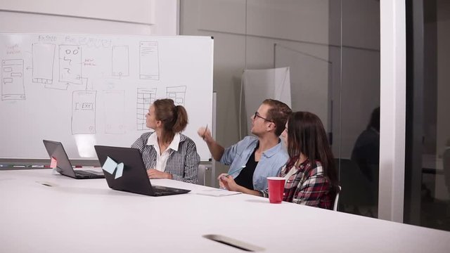 Young man in glasses and two women in casual sitting at the office table in creative workplace with laptop on it. Man in the middle pointing with pencil on whitesheet board, explaining his vision or