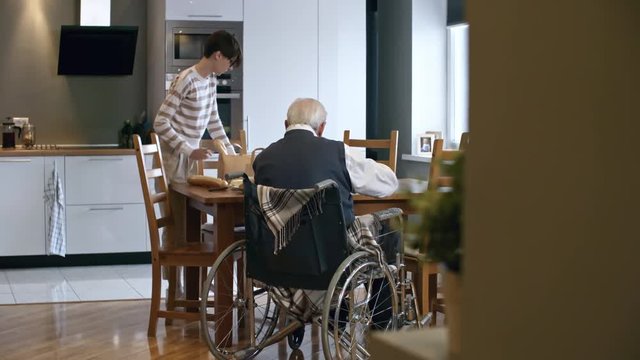 PAN of young woman in glasses unpacking groceries and talking to senior man in wheelchair sitting at dinner table